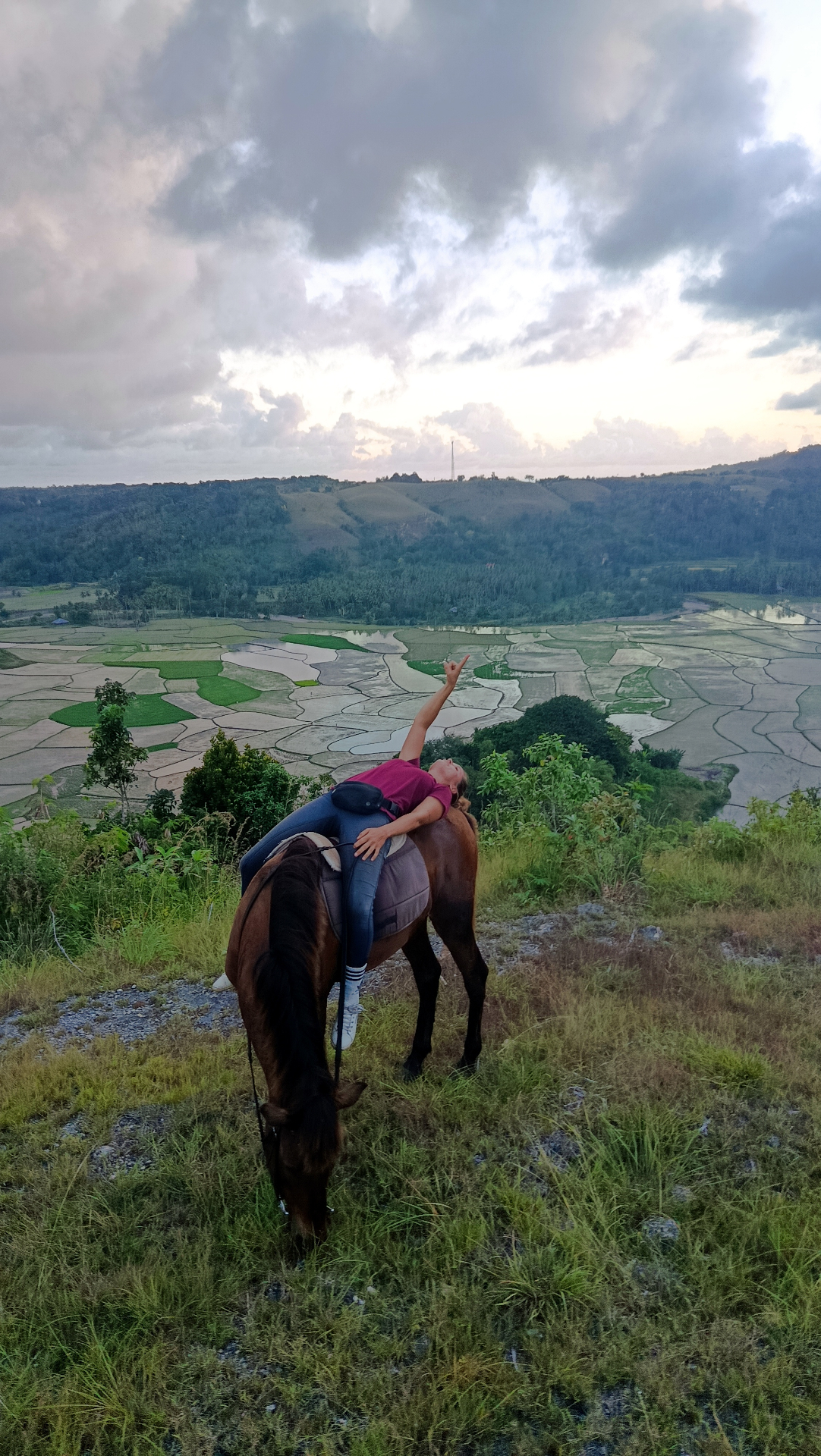 A rider lying back across a Sumba pony, pointing at the sky above rice fields at sunset